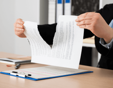 Eine Person zerreißt einen Vertrag oder ein Dokument in der Mitte über einem Schreibtisch. Auf dem Tisch befinden sich eine Klemmbrettmappe mit weiteren Unterlagen sowie Büromaterialien im Hintergrund. Die Szene deutet auf die Kündigung oder Auflösung eines Vertrages hin.