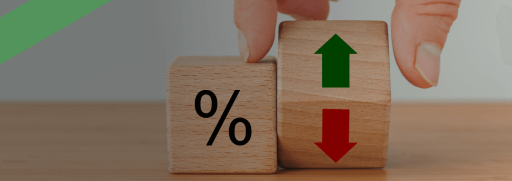 Zwei Holzklötze stehen auf einem Tisch: Einer zeigt ein Prozentzeichen, der andere grüne und rote Pfeile für Steigen und Sinken. Eine Hand bewegt einen der Klötze. Die Szene symbolisiert Zinsschwankungen und deren Einfluss auf Finanzierungs- oder Kreditkonditionen.