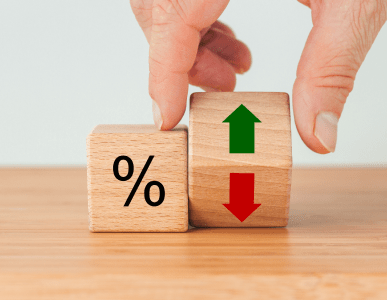 Zwei Holzklötze stehen auf einem Tisch: Einer zeigt ein Prozentzeichen, der andere grüne und rote Pfeile für Steigen und Sinken. Eine Hand bewegt einen der Klötze. Die Szene symbolisiert Zinsschwankungen und deren Einfluss auf Finanzierungs- oder Kreditkonditionen.