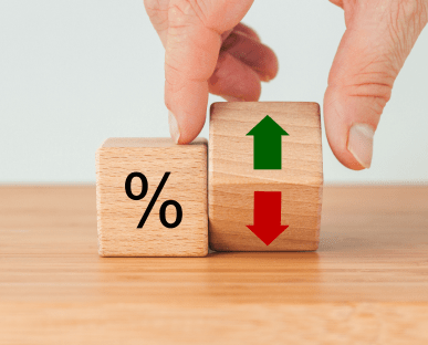 Zwei Holzklötze stehen auf einem Tisch: Einer zeigt ein Prozentzeichen, der andere grüne und rote Pfeile für Steigen und Sinken. Eine Hand bewegt einen der Klötze. Die Szene symbolisiert Zinsschwankungen und deren Einfluss auf Finanzierungs- oder Kreditkonditionen.