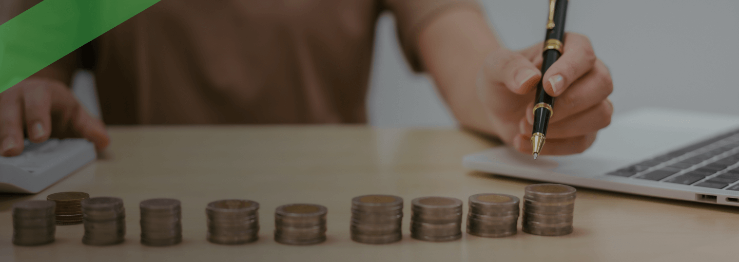 Eine Person sitzt an einem Schreibtisch und arbeitet mit Taschenrechner und Stift. Vor ihr sind mehrere gleichmäßig gestapelte Münzstapel aufgereiht. Die Szene symbolisiert finanzielle Planung und Kalkulation, etwa im Zusammenhang mit einer Zinsbindungsfrist bei Krediten.